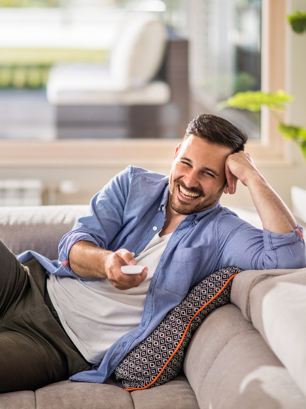 Young cheerful man relaxing on sofa in the living room and changing channels with remote control.